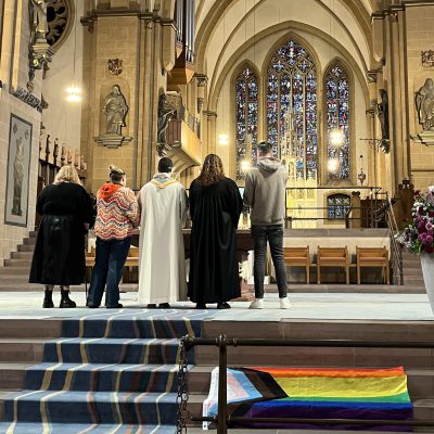 5 Personen stehen in einer Kirche vor dem Alter. Auf der Treppe davor liegt eine Regenbogenflagge
