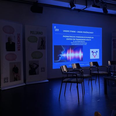 Projektion einer Präsentation über stimmangleichende Maßnahmen bei trans* Personen. Davor stehen Stühle für eine Podiumsdiskussion.