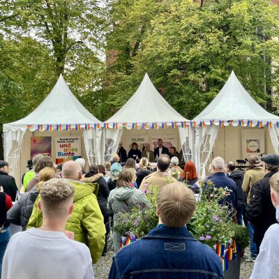 Menschenmenge vor Festzelten, die mit Regenbogenfahnen geschmückt sind